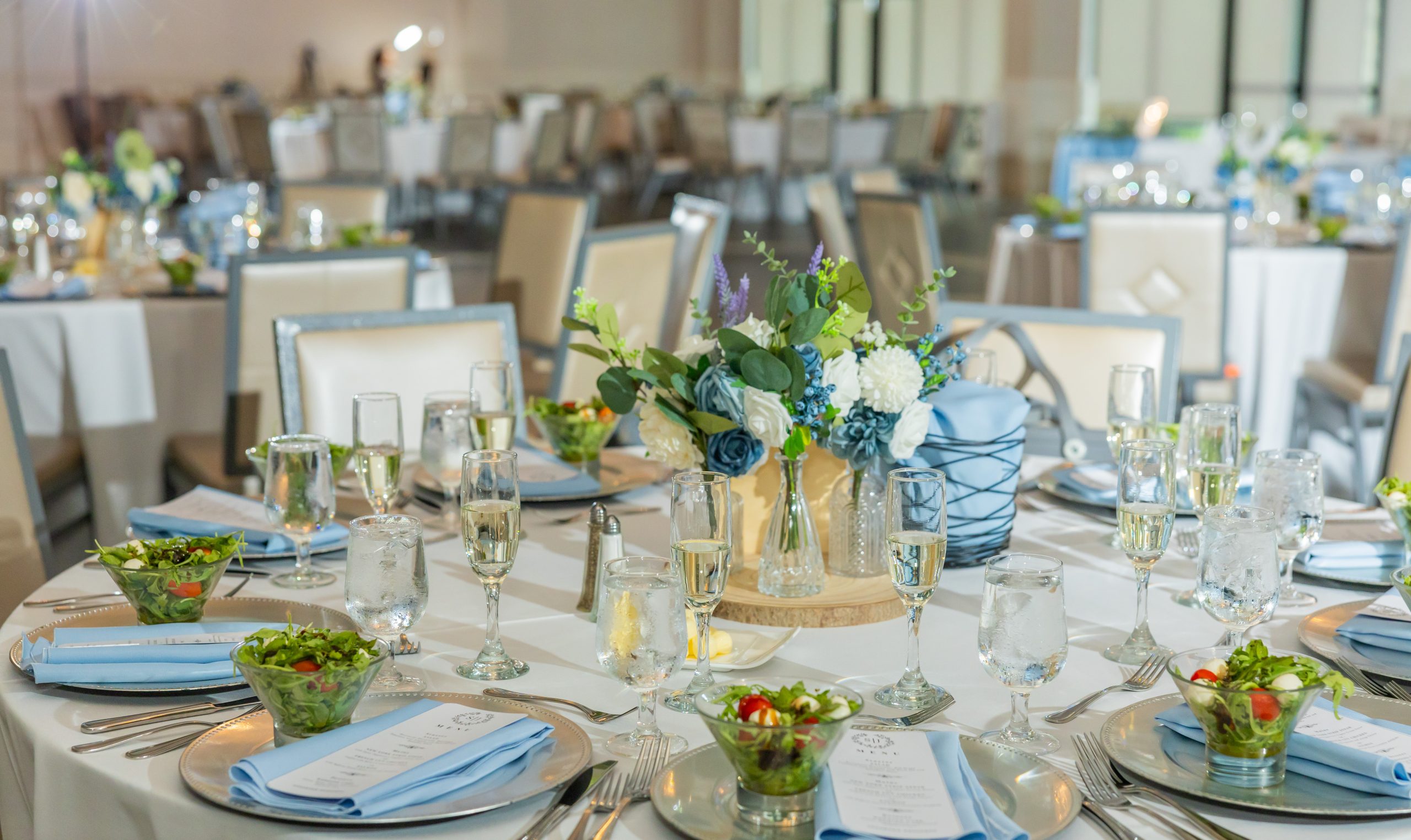 The ballroom set for dinner service at The Clubhouse at Galloping Hill in Kenilworth, New Jersey with blue napkins, silver chargers, salads and champagne glasses.