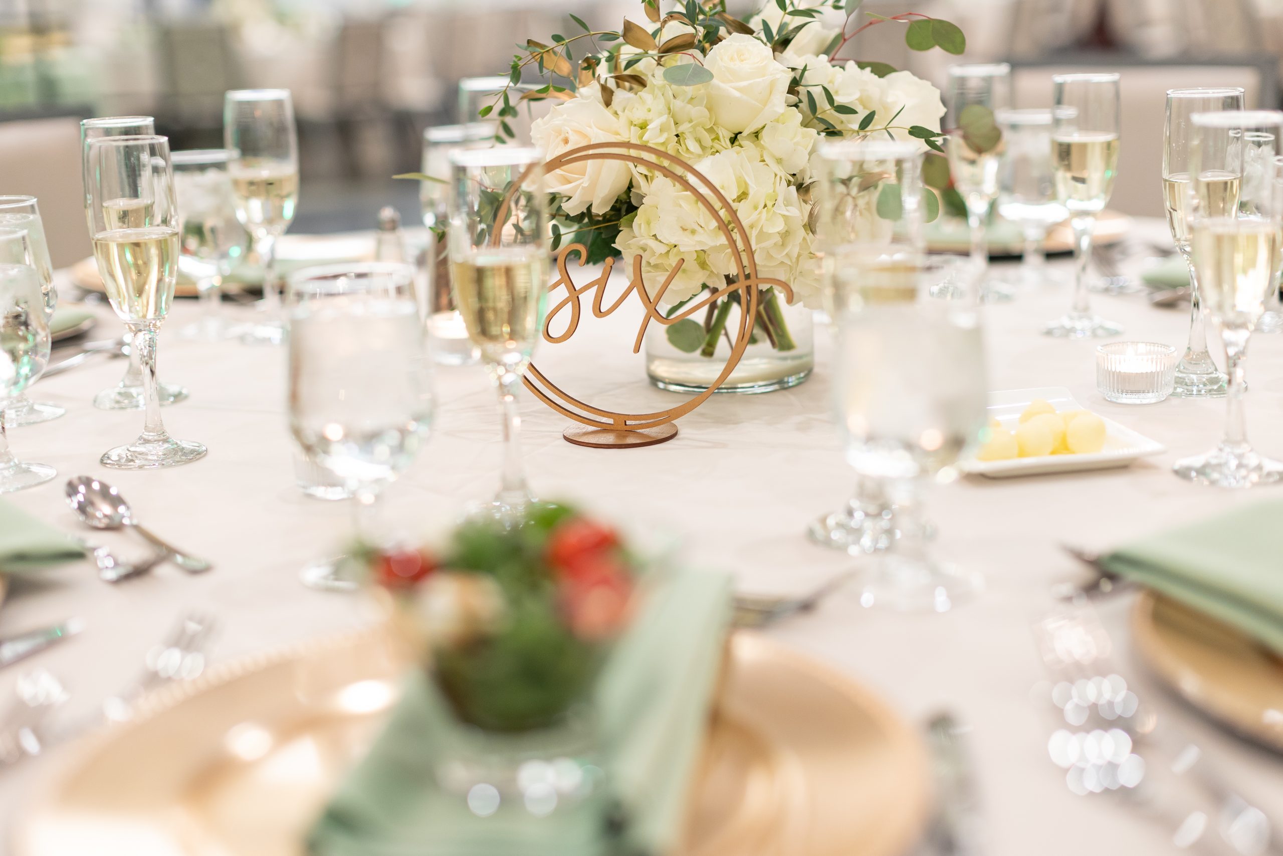 A table set for a service with gold chargers, green linen, salads, champagne glasses, and a table number at The Clubhouse at Galloping Hill in Kenilworth, New Jersey