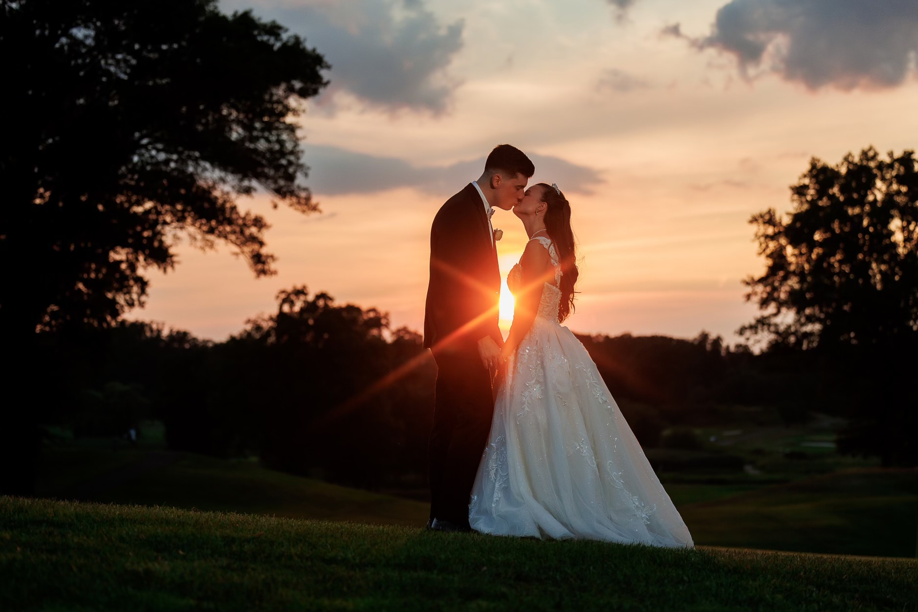 A bride and groom kissing in front of a sunset at The Clubhouse at Galloping Hill in Kenilworth, New Jersey