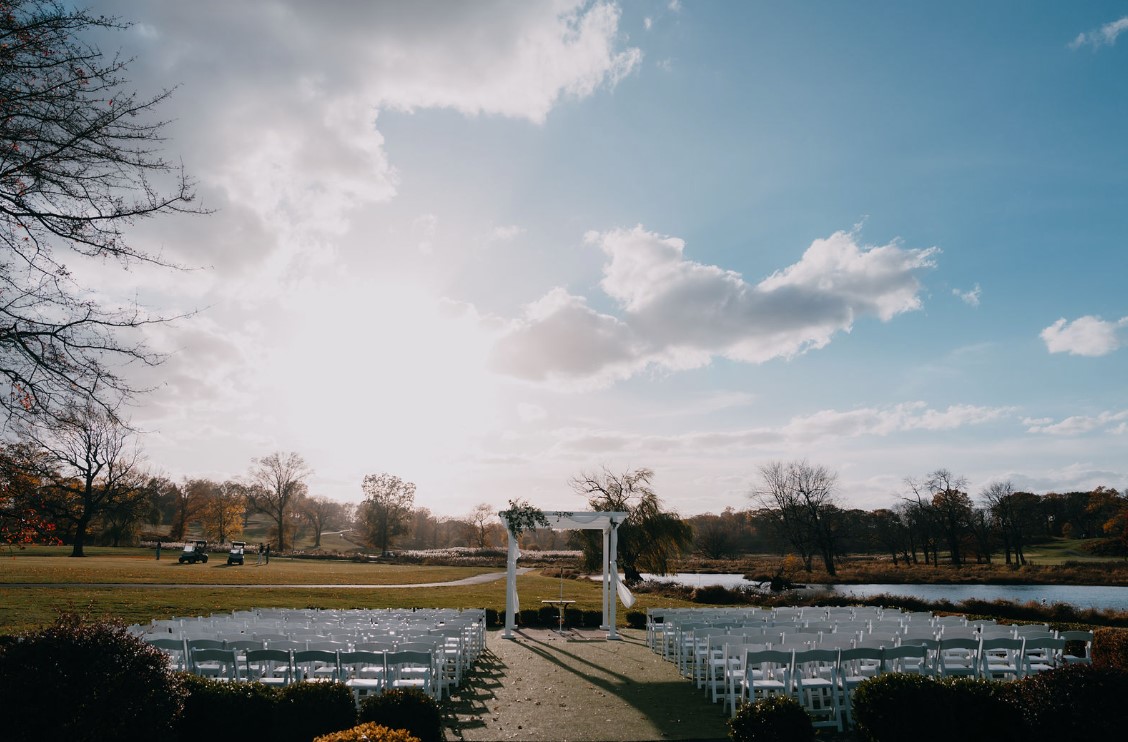 The outdoor ceremony site at The Clubhouse at Galloping Hill in Kenilworth, New Jersey, set up with white folding chairs in two columns, golf carts and a pond in the background
